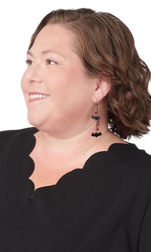 A women wearing dangling Halloween earrings featuring two small black bats separated by small, faceted red beads on a thin chain.