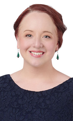 A smiling woman wears green, elegant dangling earrings shaped like Christmas trees.