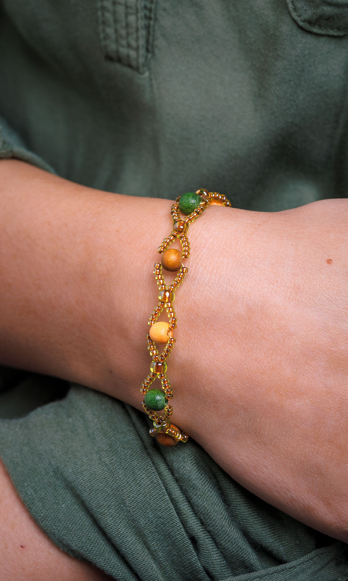 woman's wrist over other arm wearing Design Idea S B 3 6 &ndash; Bracelet features dark red-, green-, and honey-hued wooden beads surrounded by gold-colored seed beads.