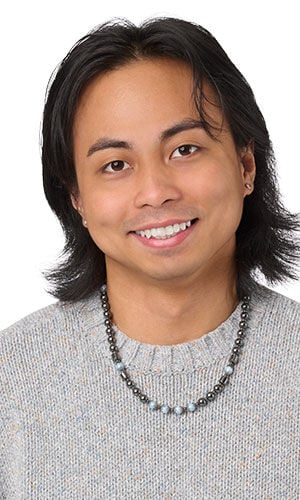 A beaded necklace of dark metallic and light gray stones, worn by a smiling man in a gray sweater.