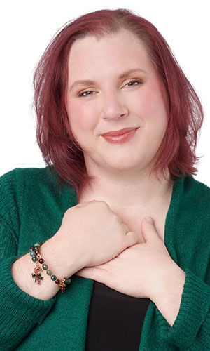 Smiling woman with shoulder-length reddish-purple hair and a green sweater holds their hands to their chest, displaying a beaded charm bracelet