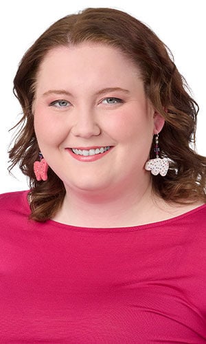 Model in a pink shirt wearing design idea S B 2 U pink and white polymer clay animal cracker earrings with colorful sprinkle details, accented by faceted purple crystal beads and finished with silver ear wires.