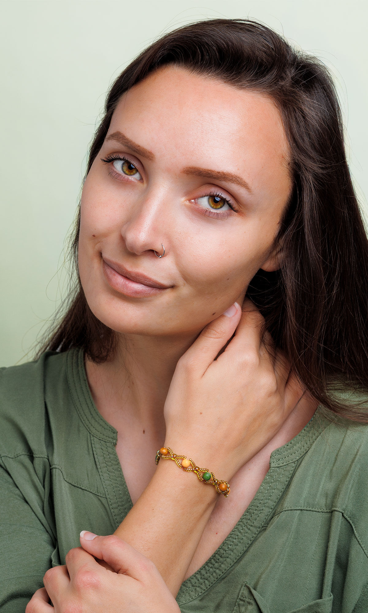 woman in a green top with hand on her neck wearing Design Idea S B 3 6 &ndash; Bracelet features dark red-, green-, and honey-hued wooden beads surrounded by gold-colored seed beads.