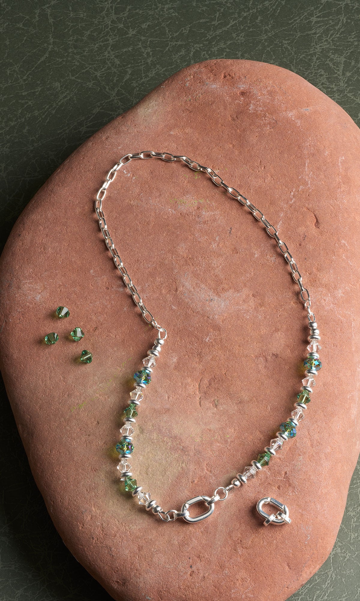 A silver chain necklace with green and clear crystal beads resting on a large reddish-brown stone.