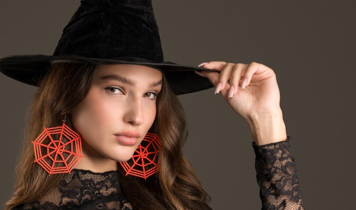 Model wearing a black hat and black clothing wearing large earrings made from orange seed beads and designed to look like spiders’ webs