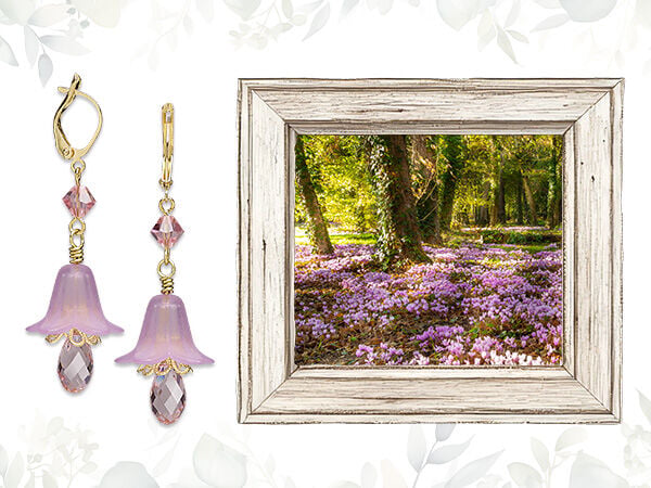On the right, a gray wood frame surrounds an image of a field of light purple flowers in a forest. On the left is a pair of golden earrings, each with a downward-pointing light purple flower bead with a light purple faceted briolette drop below it.