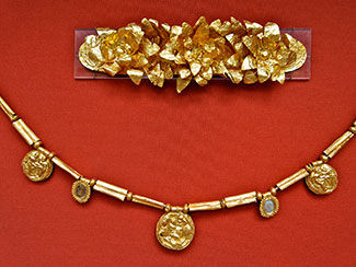 Ancient gold jewelry consists of a brooch with petaled flowers and a chain with long gold beads and gold coin-shaped pendants.