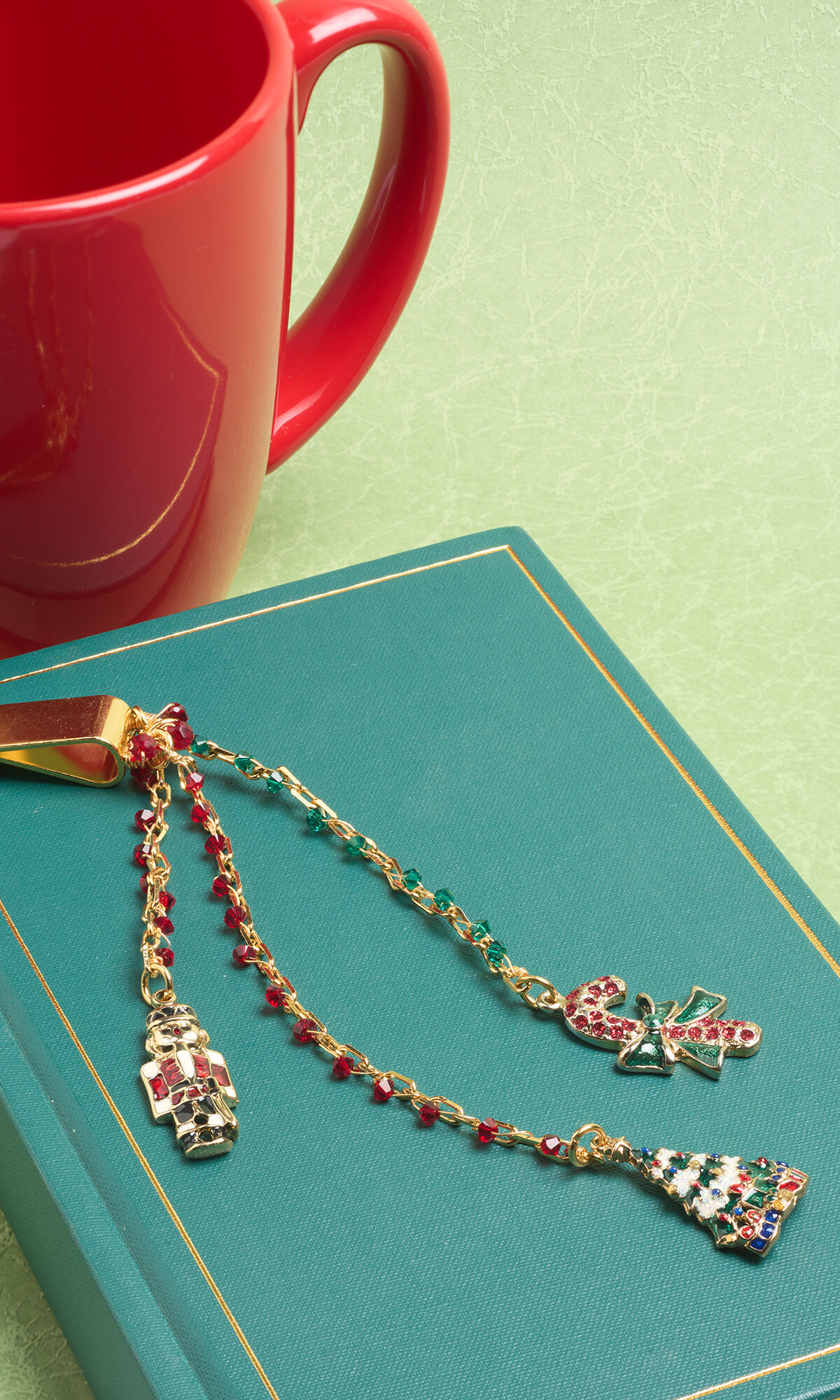 A holiday-themed metal bookmark clip with three gold chain dangles rests on a green book. The chains are accented with small red and green beads and feature festive charms: a nutcracker, a candy cane with a bow, and a decorated Christmas tree. A red mug sits in the background.