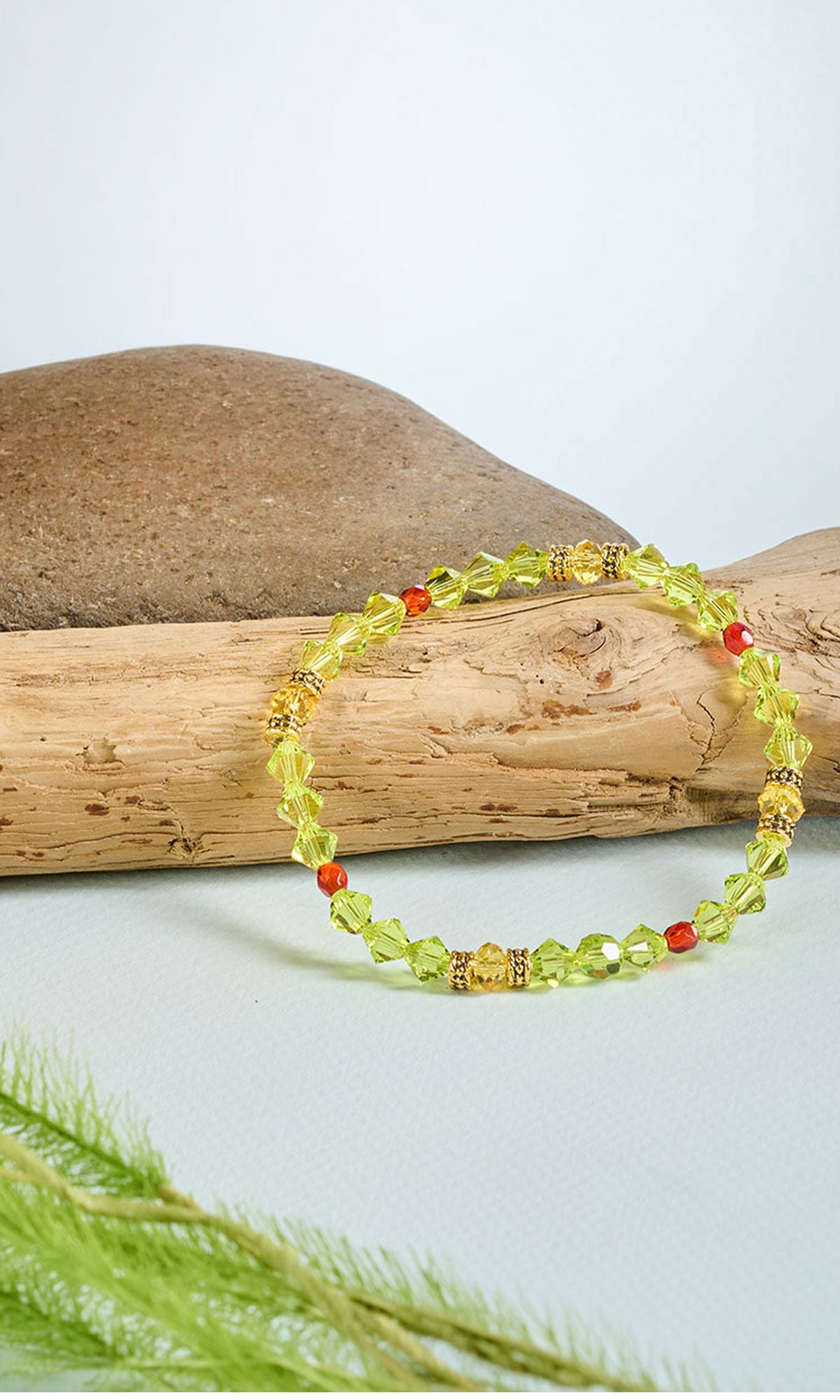 citrus green and red bracelet place on a wooden stick with a rock behind on a light blue background