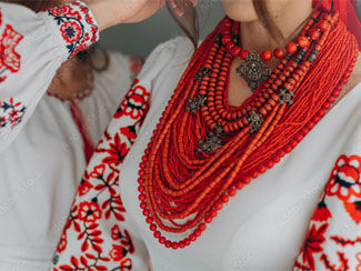 Woman in white dress with coral-hued embroidery on the sleeves wearing dozens of strands of coral and metal necklaces.