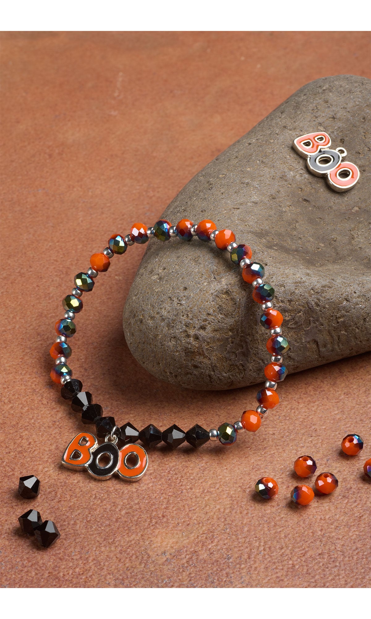 A beaded Halloween bracelet with orange, black, and iridescent beads, featuring an orange and silver "BOO" charm, displayed on a brown surface next to a grey rock and loose beads.