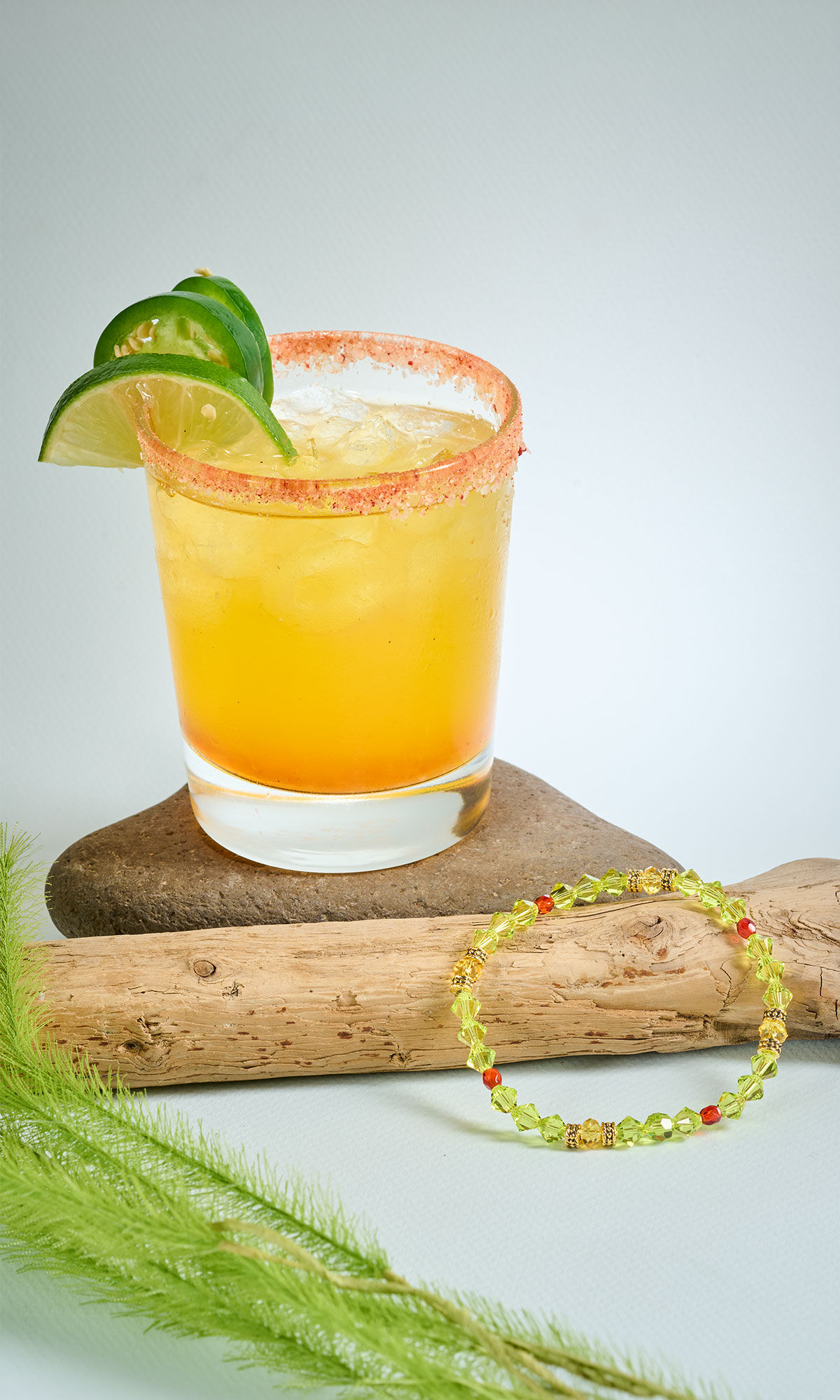 Glass with orange liquid garnished with limes. A green and red bead bracelet rests against a piece of driftwood nearby