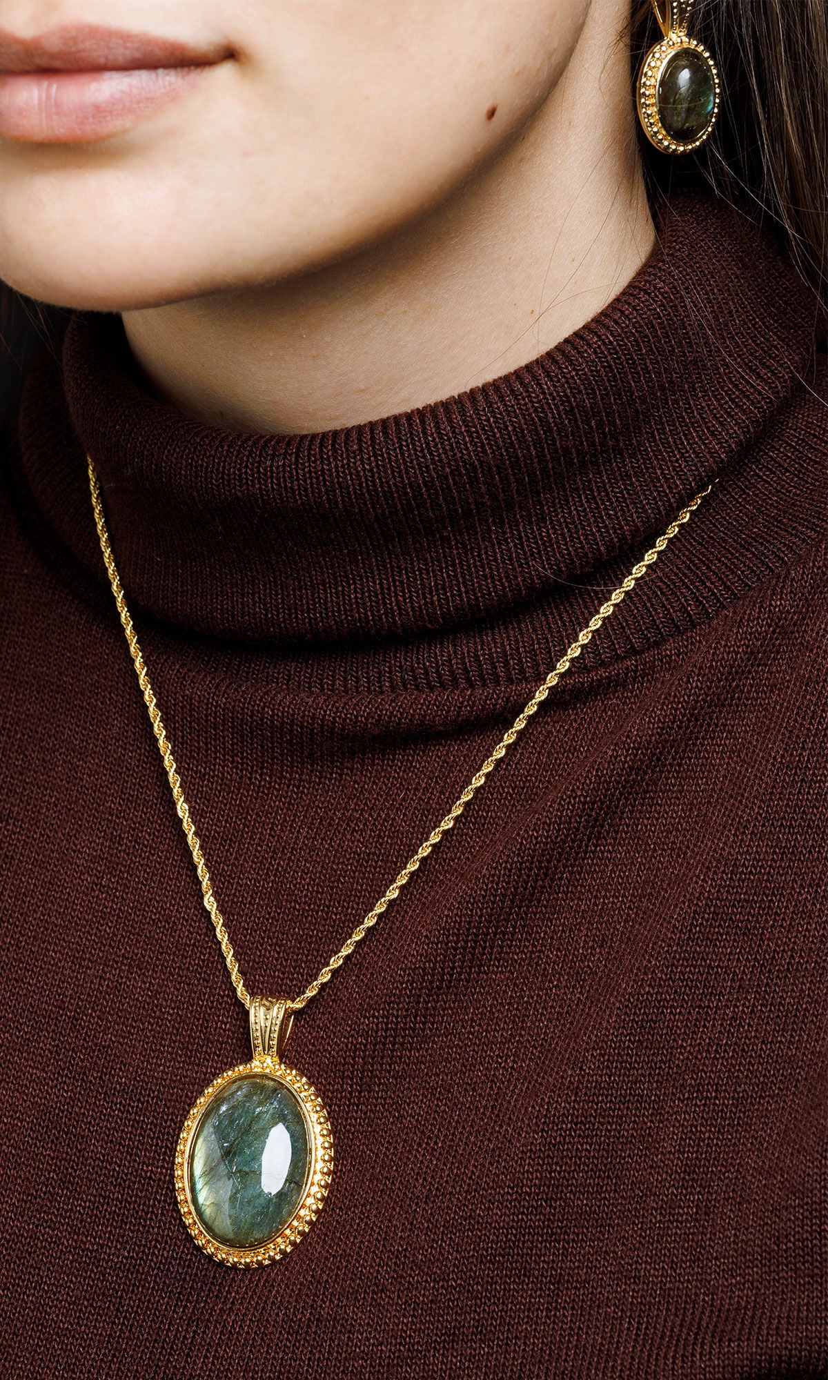 A woman wearing a brown turtleneck and gold jewelry with oval green labradorite gemstones.