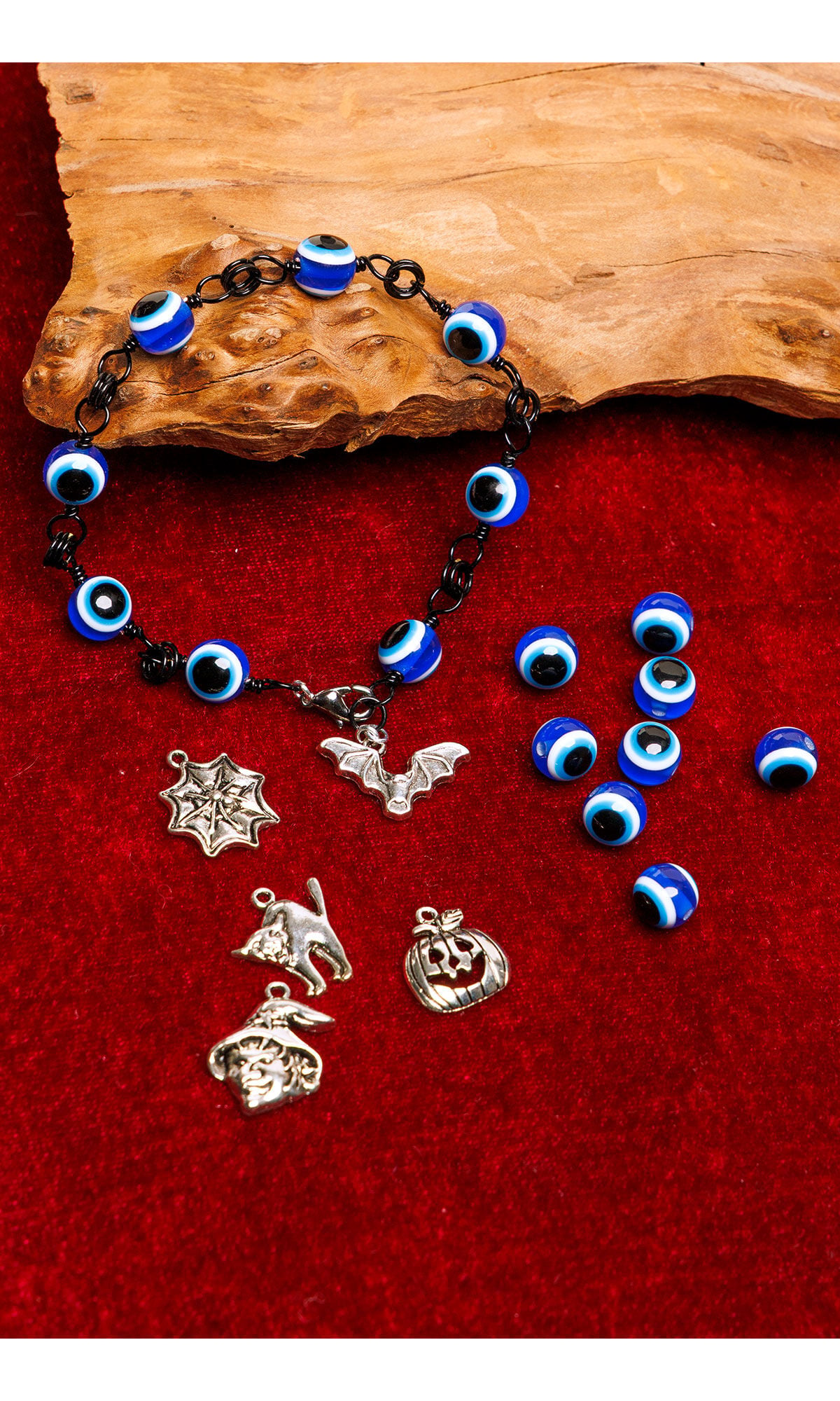 A bracelet with blue eye beads and metal charms lies on a red background. Nearby are loose beads and pewter charms, including a bat, witch, spider in web, cat and a pumpkin.