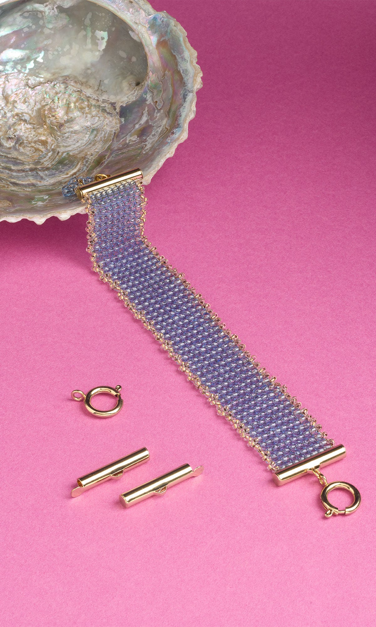 A wide beaded bracelet made of small, light blue beads with a gold trim lies flat on a pink surface. One end of the bracelet is tucked inside a large, iridescent seashell. Loose gold-toned jewelry findings, including a round spring clasp and two slide end tubes, rest on the surface next to the bracelet.