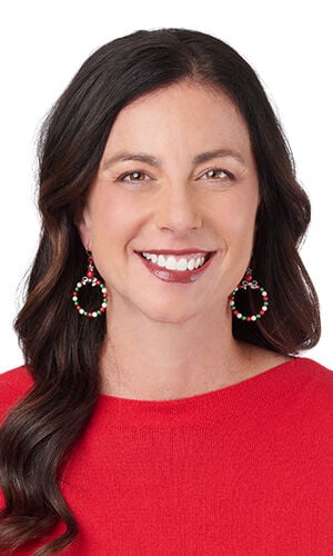 A woman with brown hair smiles, wearing circular Christmas wreath earrings made of red, white, and green beads.
