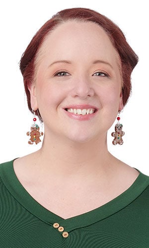 A smiling person with auburn hair tied back and a green v-neck top wears festive gingerbread man earrings