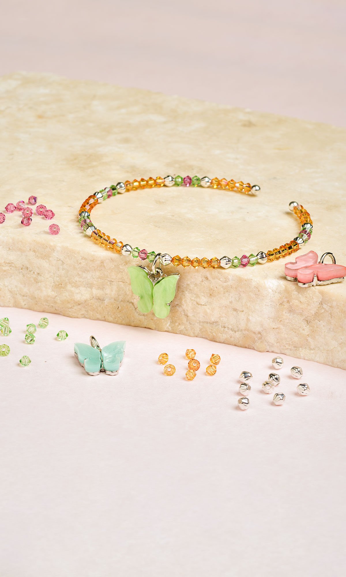 Design idea R J 2 N is a beaded memory-wire bracelet with faceted pink, green and orange crystals, silver-plated beads and an iridescent butterfly charm laying on a slap of light beige granite