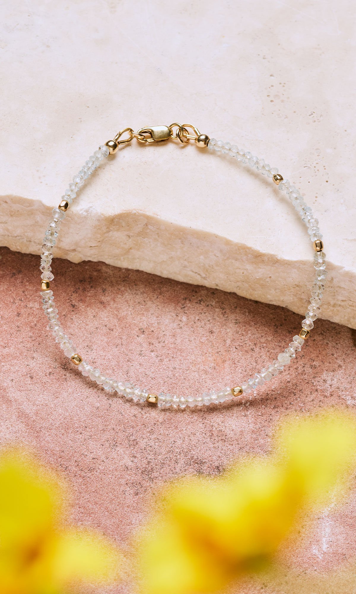 Design Idea R G 2 J Bracelet composed of many small white faceted gemstones interspersed with even smaller gold beads laying on a piece of beige marble with blurred yellow flowers in the foreground
