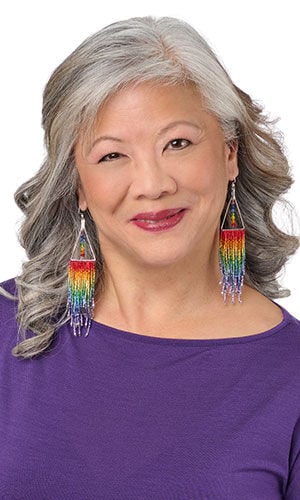 Woman wearing large, prominent triangular silver earrings with long, colorful rainbow-beaded fringe.