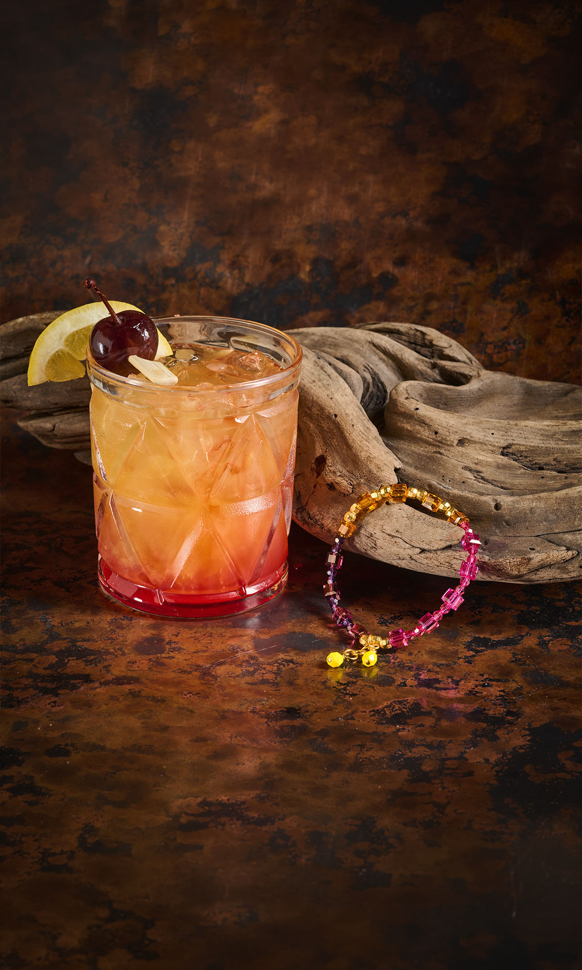 Glass with pink and yellow liquid inside with a purple and gold bracelet resting on driftwood nearby