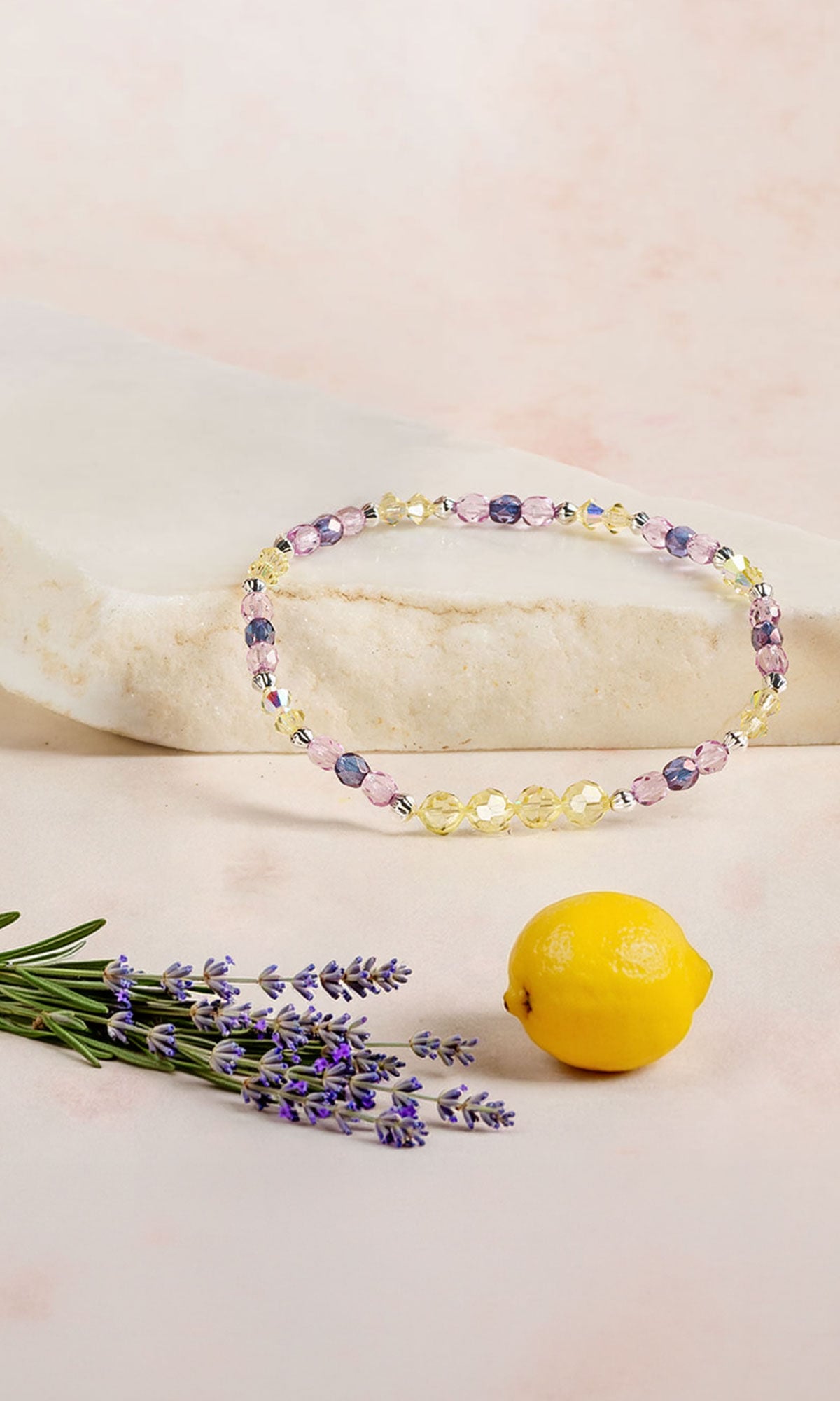 Lavender and Jonquil yellow bracelet lying on a piece of light beige granite with a lavender flower stem and a small lemon in front.