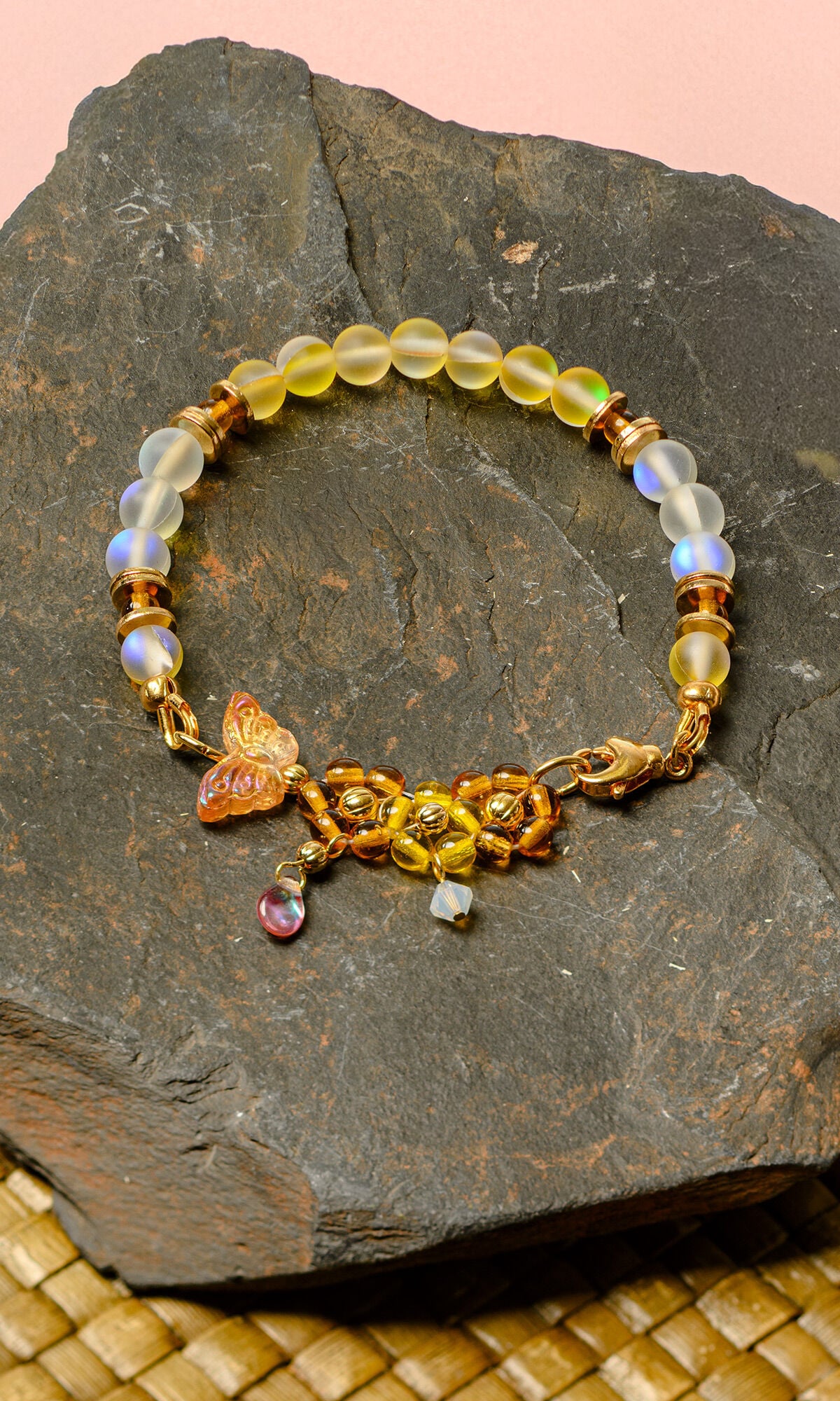Design Idea S B 2 N &ndash; Bracelet features iridescent white and yellow glass beads, yellow and orange glass beads configured to look like flowers, and a gold-colored glass butterfly bead. It is sitting on a large, flat rock on top of a tan woven material