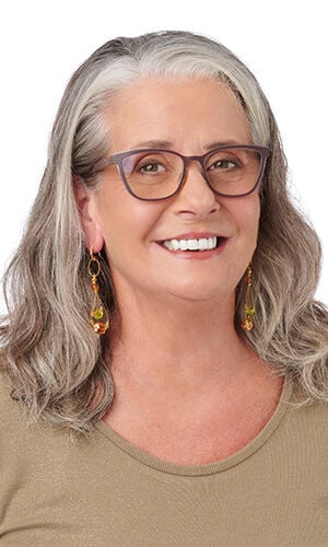 A smiling woman wearing a tan top and long, gold-toned dangle earrings featuring amber and green beads..