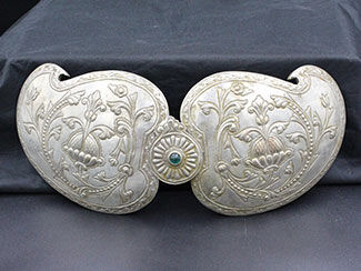Silver buckle has paisley-shaped components on either side featuring foliage and flowers. Central component has a blue stone and a flower design.