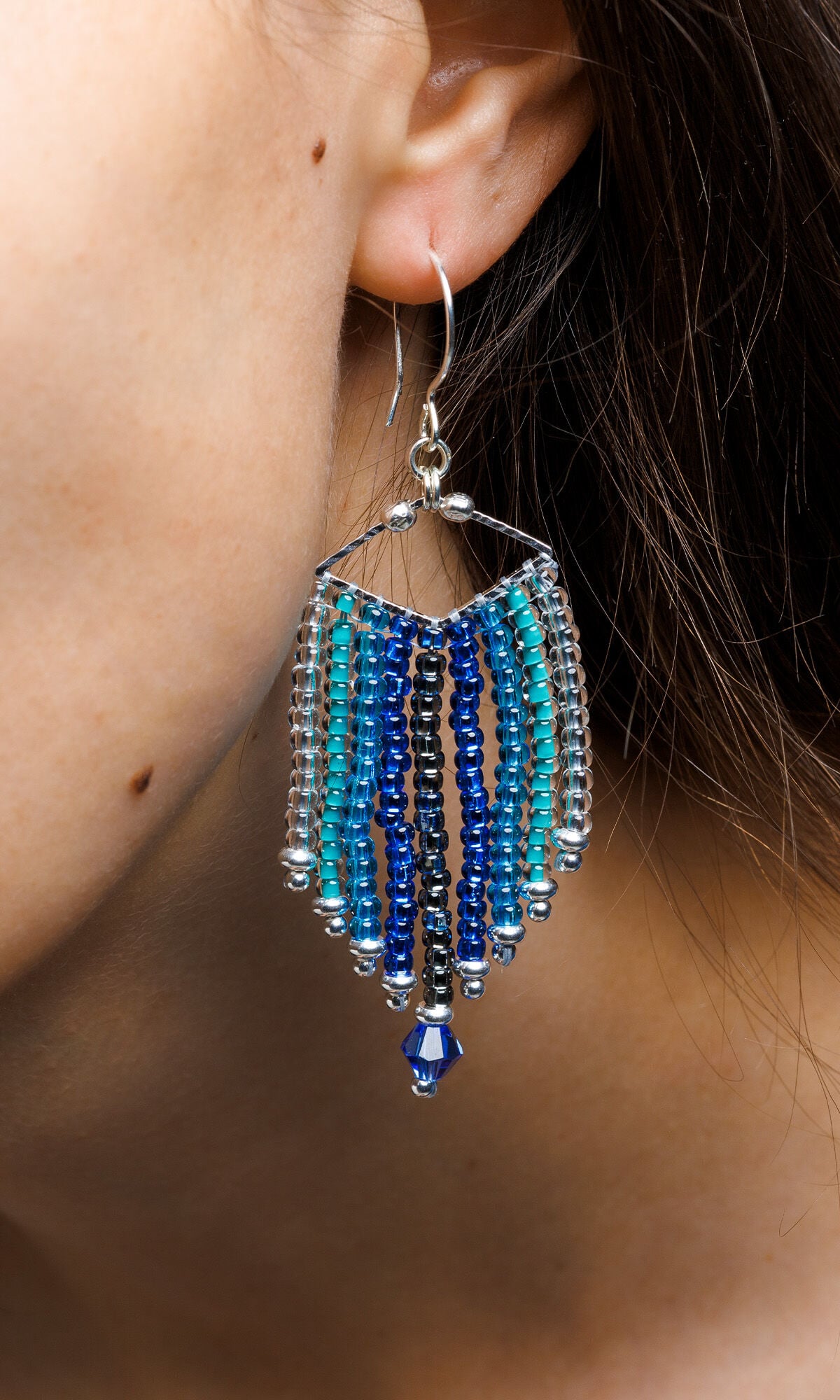 Closeup of ear wearing Design Idea S B 2 X &ndash; Each earring has a silver diamond wider than it is tall with nine columns of seed beads hanging from it in various shades of blue.