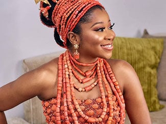 Her head is styled and crowned with coral beads which form a crown on her head. Also wearing several coral bead necklaces. 