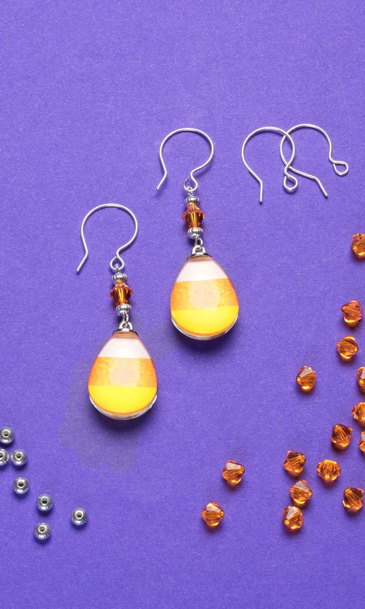 Close-up of candy corn earrings on a purple background, surrounded by small orange and silver beads. Bright and playful vibe.