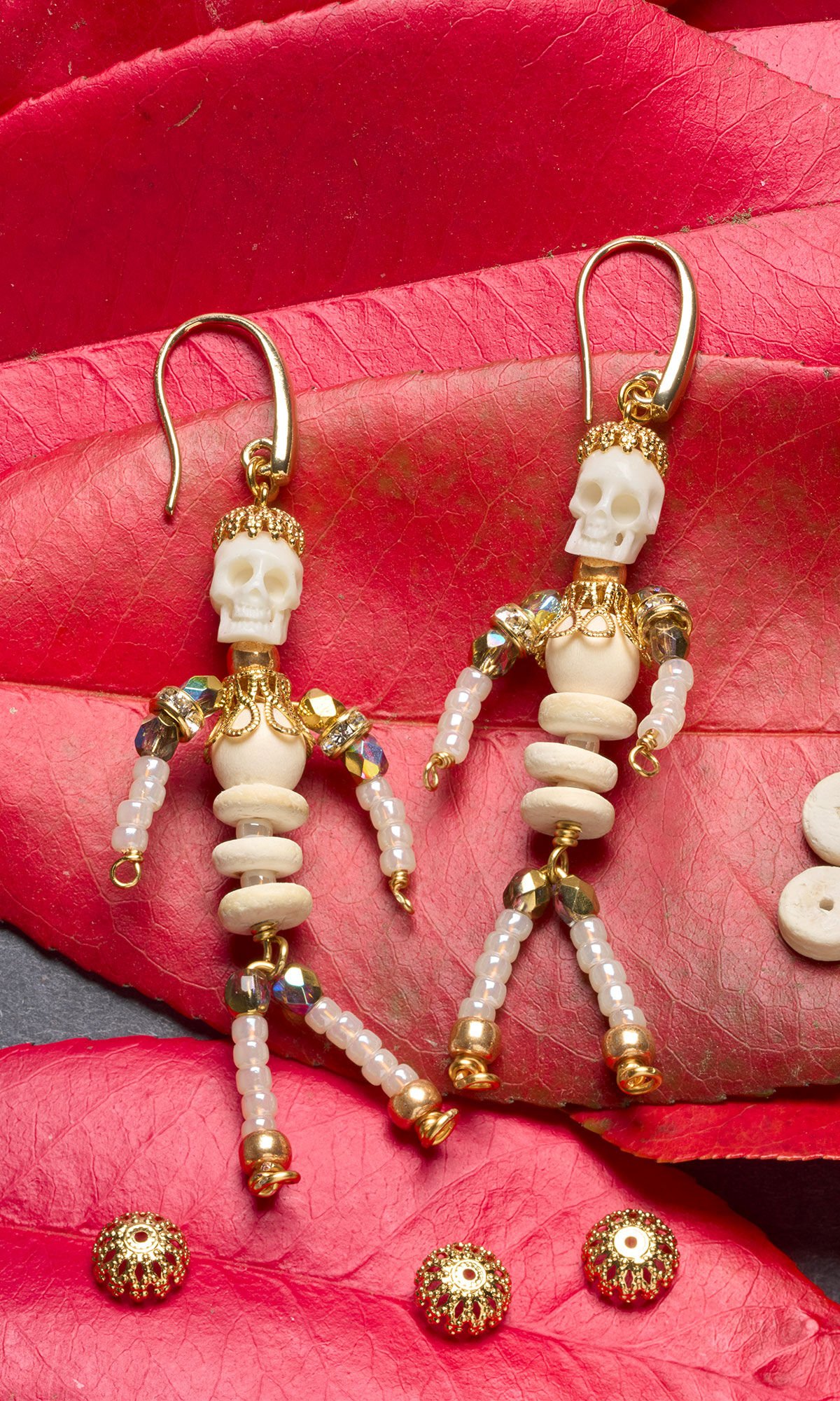 A pair of skull earrings with beaded, articulating skeleton bodies resting on a bright red leaves.