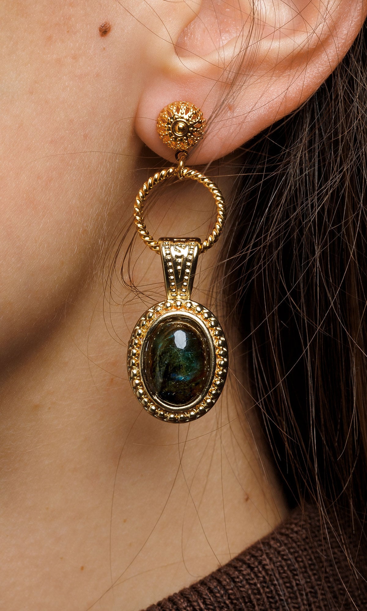 A close-up of a person's ear wearing a large, ornate gold drop earring featuring a dark, iridescent oval gemstone.
