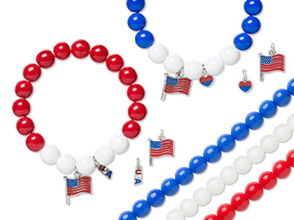 Two beaded bracelets, one white and red and the other white and blue, are at the top of the image. Both have American flag and &ldquo;U S A&rdquo; charms. Three strands of round, shiny beads lie diagonally across the bottom right corner: blue, white and red.