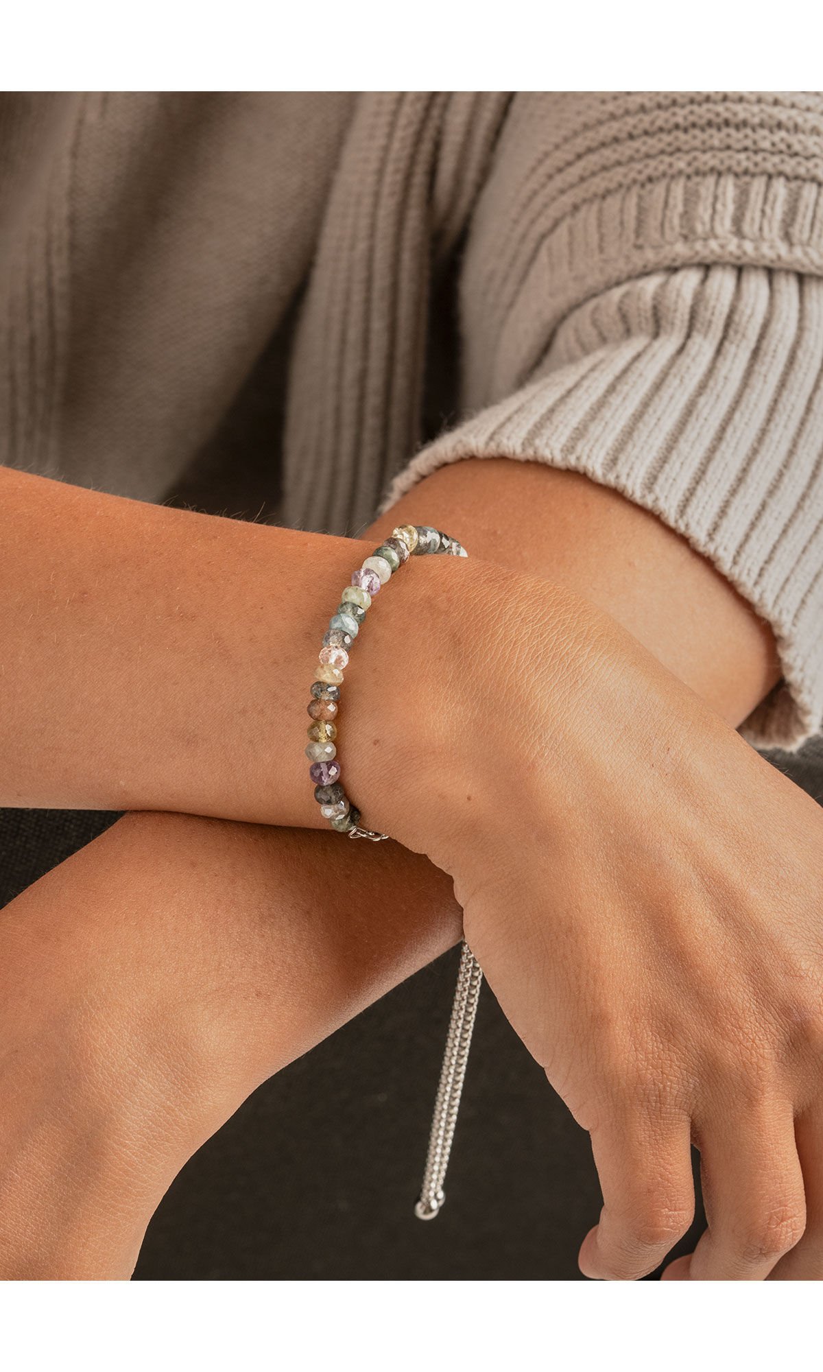 close up shot of model's wrist wearing design idea R G 2 Y Bracelet.  Half of the bracelet is subdued shades of multi-colored gemstone beads. The other half is a thick chain that ends in two steel balls and has a large slider bead to adjust size.