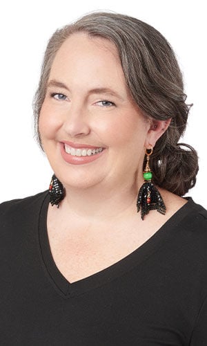 Woman wearing mock witch earrings, styled as large dangle earrings with a vibrant green bead and a flared black beaded mesh tassel resembling a whimsical witch's hat or skirt.