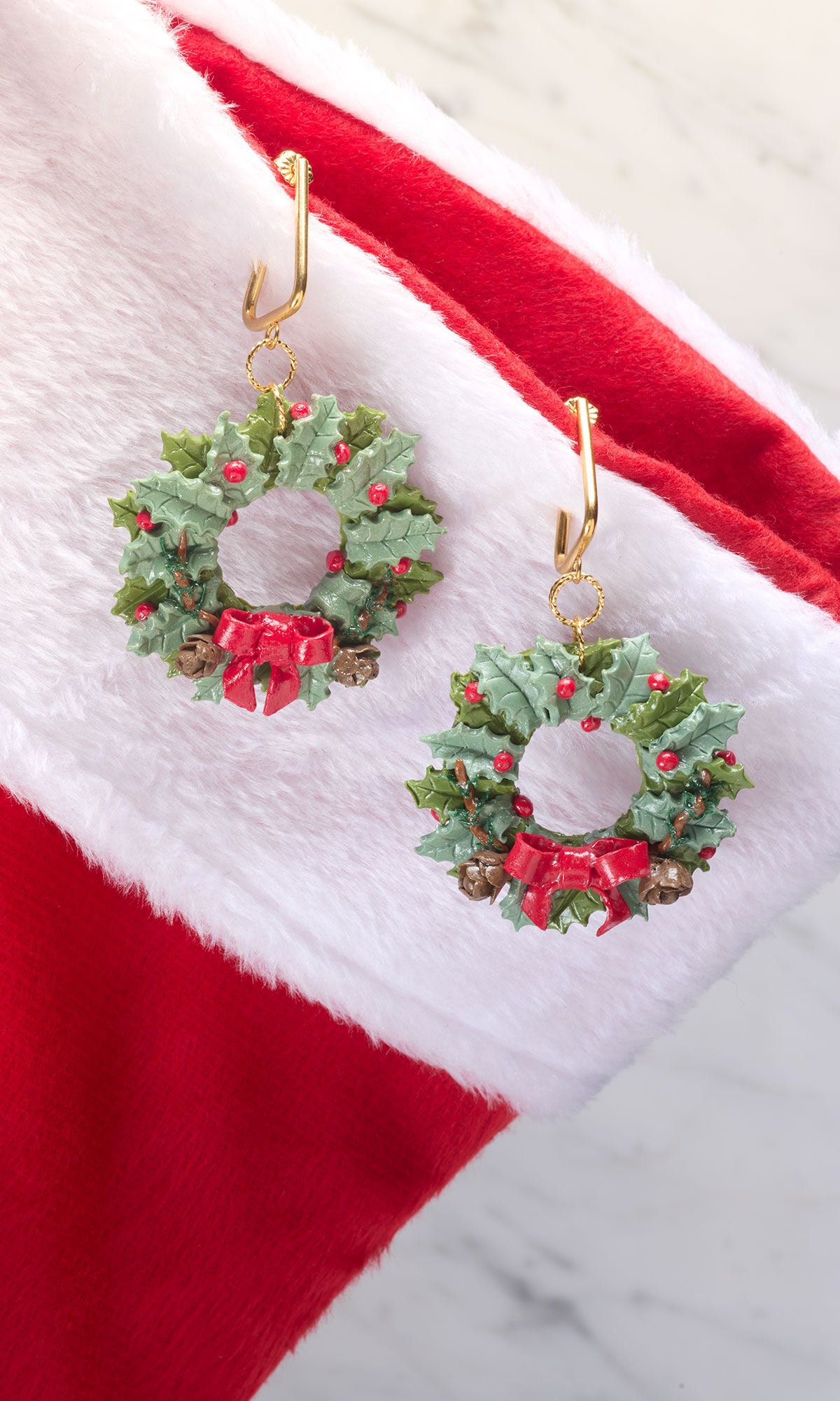 A pair of festive Christmas wreath earrings, featuring detailed green holly leaves, small red berries, and a bright red bow, hanging from gold hooks against a white and red fuzzy holiday stocking.