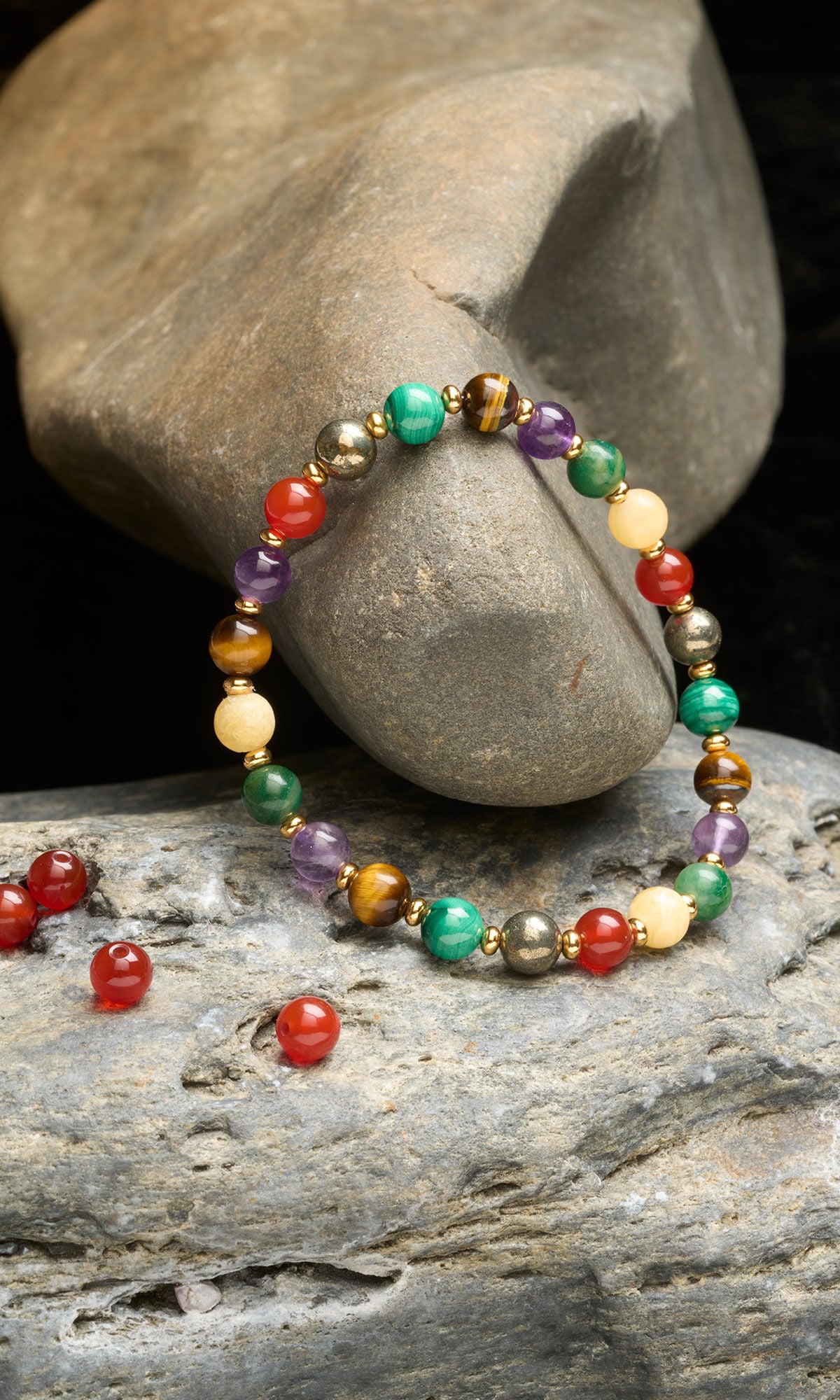A multi-colored beaded bracelet featuring gemstones like tiger's eye, malachite, amethyst, and carnelian with small gold spacers, displayed on natural gray stones.