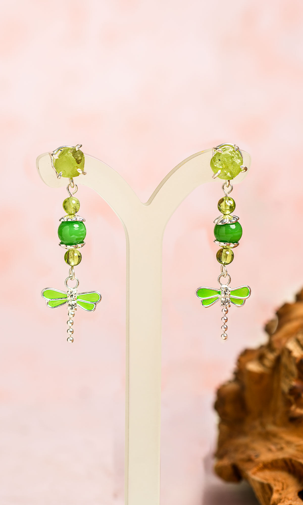 Silver-toned drop earrings with green beads and dragonfly charms on a white stand against a soft pink background.