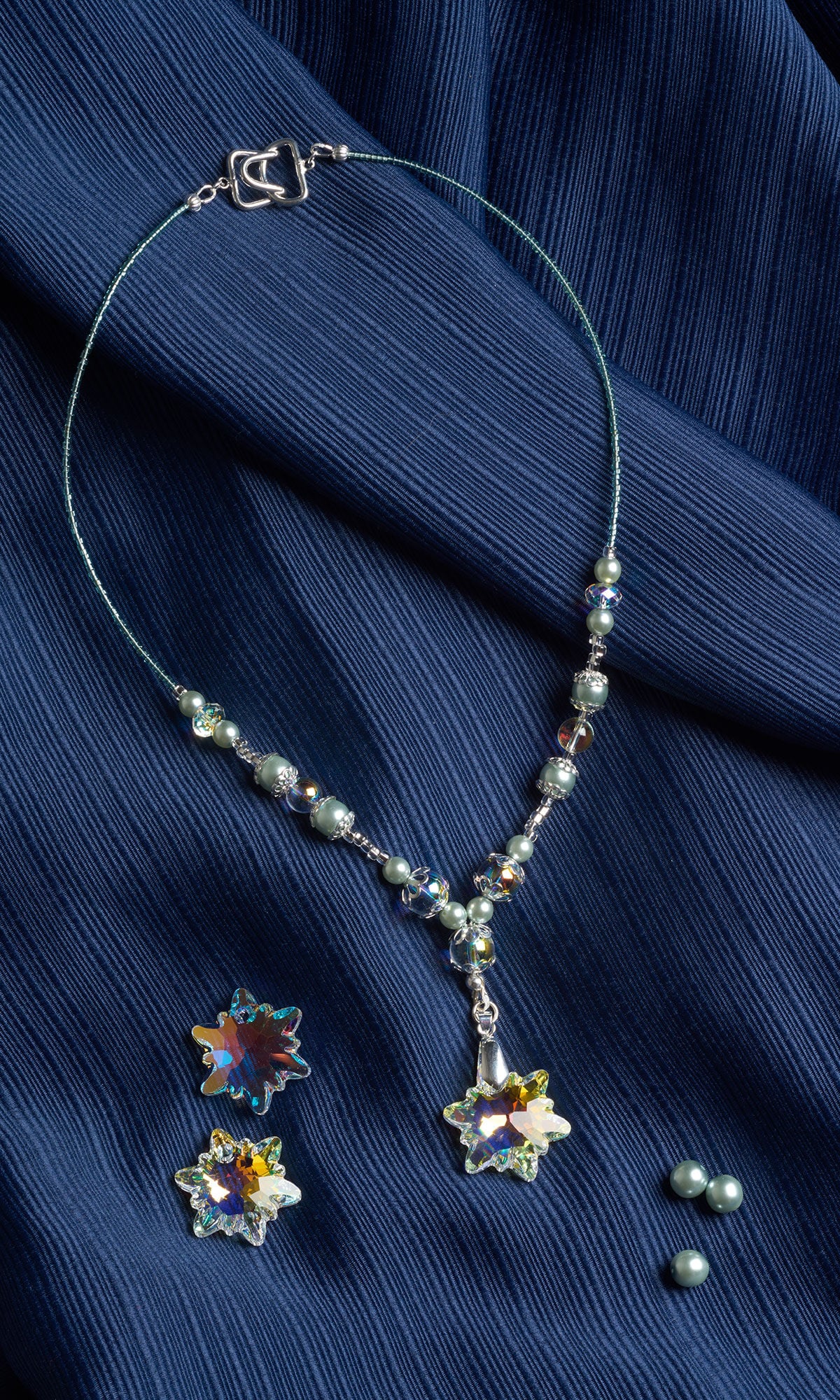 A silver-toned crystal necklace and matching earrings featuring star-shaped irised pendants are displayed against a textured dark blue fabric background.