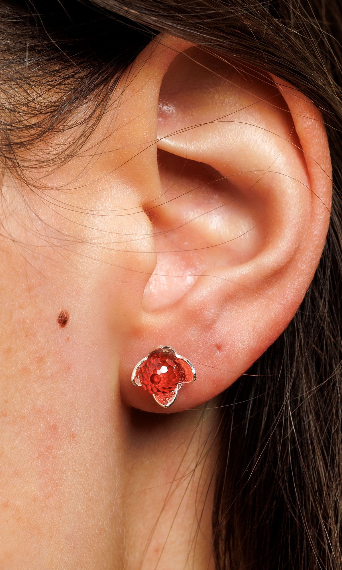 Close-up of an ear wearing a silver flower-shaped earring with a vivid red crystal in the center.