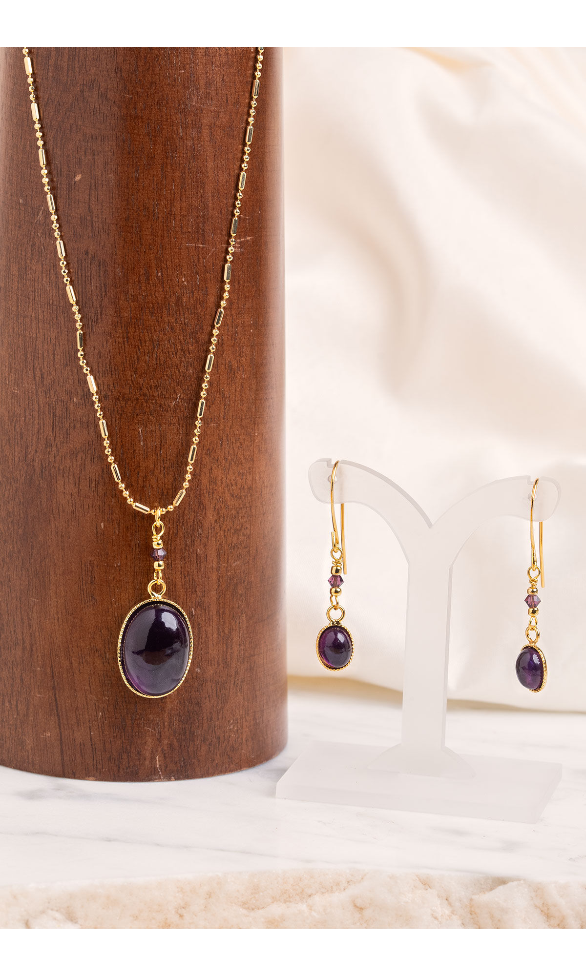 Design Idea R K 2 F - Gold necklace and earrings feature simple amethyst oval cabochons beneath short, beaded strands against a wood and white fabric background.
