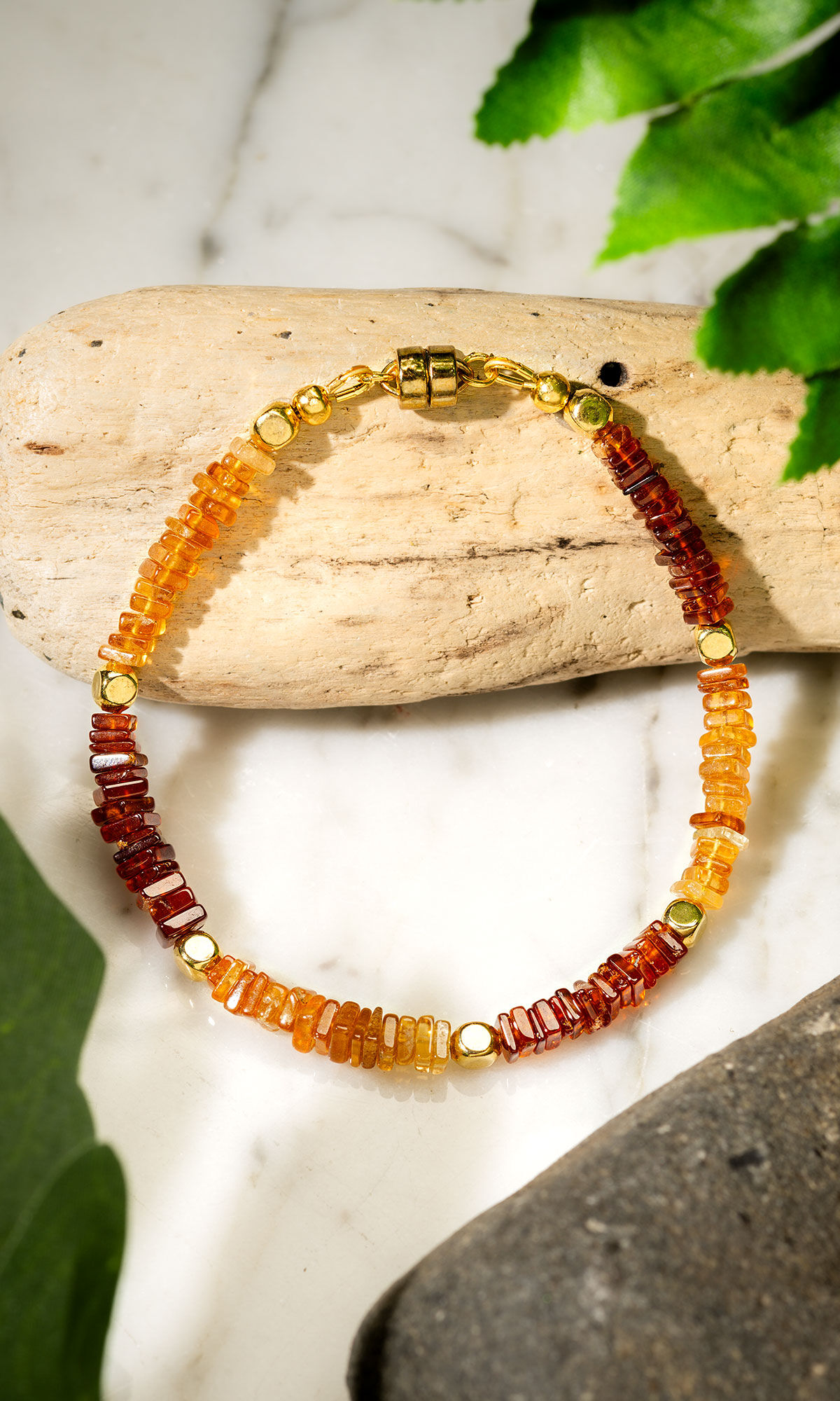 Design Idea R G 2 M - Single-strand bracelet with dark and light honey-toned garnet beads separated by gold-plated square beads laying on top of a piece of wood the green foliage and a rock