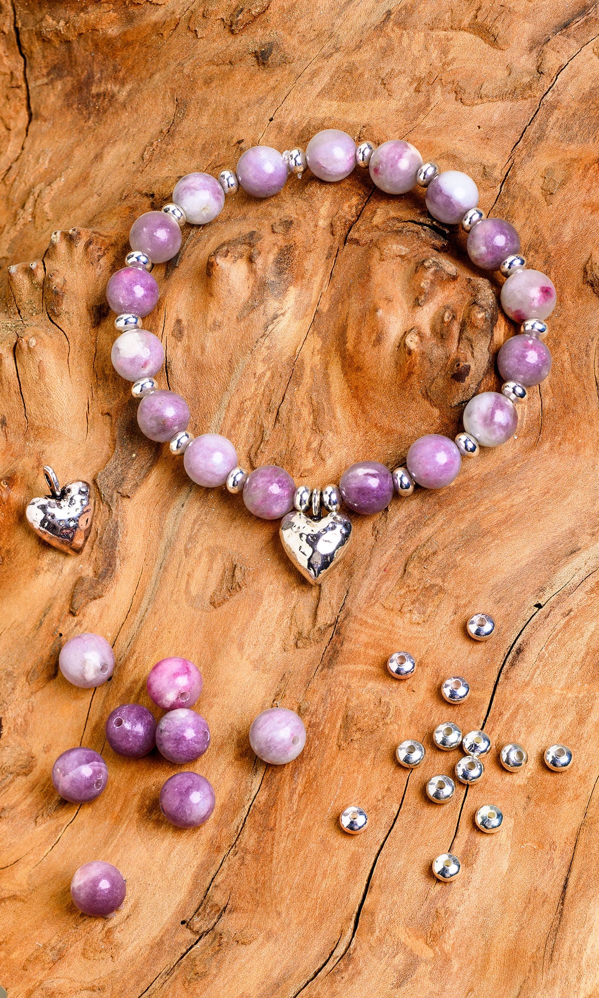 Purple gemstone bracelet with a silver heart charm laying on a rustic wood background. Loose purple beads, a second small heart charm, and several silver spacer beads are scattered around the main bracelet.