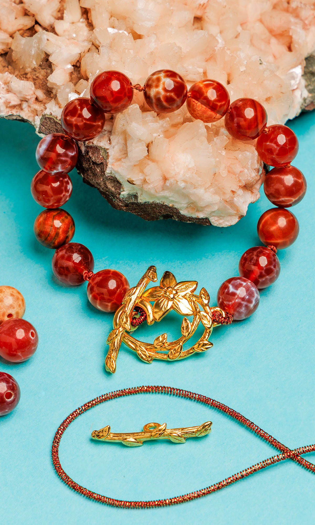 A beaded red agate bracelet with a gold floral toggle clasp, displayed on a teal surface alongside raw crystals and jewelry-making components.