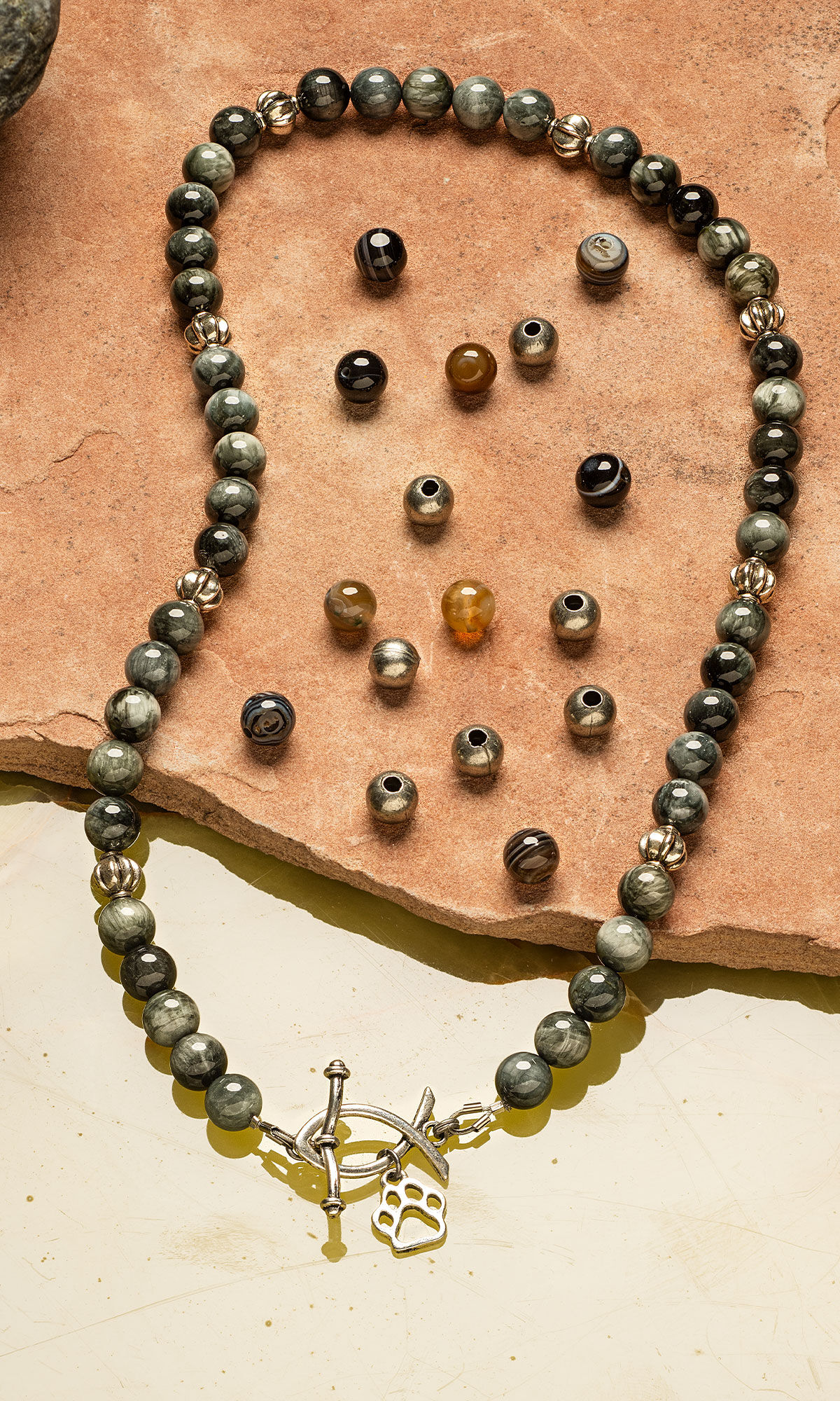 Necklace of glossy grey-toned beads with a silver front clasp in the shape of a Christian fish and a paw print charm on a terracotta stone and beads spread out