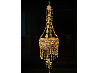 Cylindrical gold &ldquo;crown&rdquo; has blue and white gemstones. A cross hangs down from the center, and gold drops hang from the edges with alphabet letters and gemstones