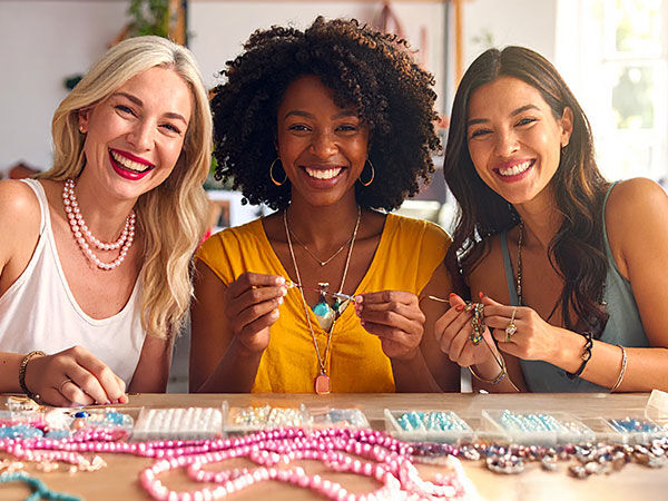 3 young woman between the ages of 30-40 with smiling faces sitting at a workbench make jewelry 