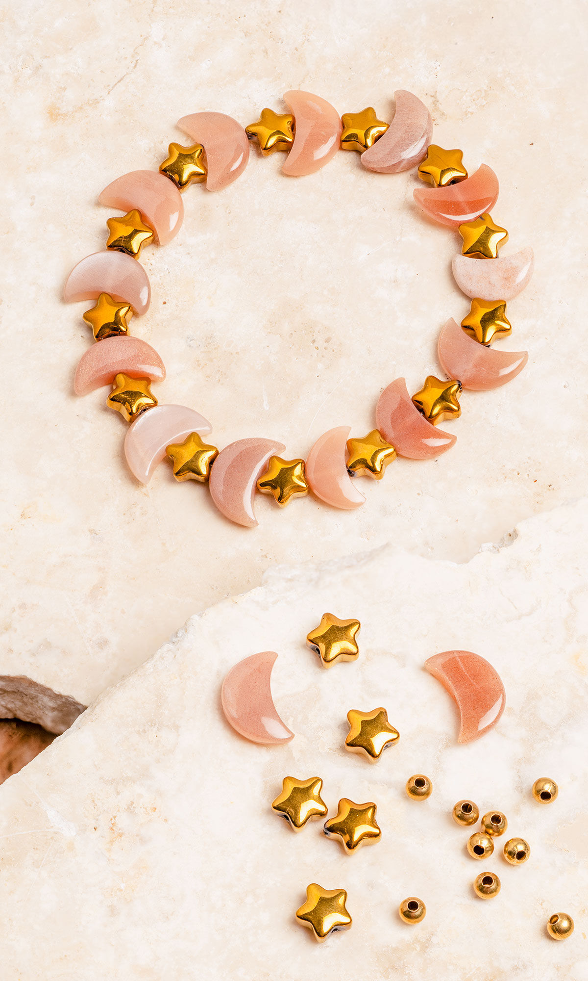 A beaded bracelet featuring alternating peach-colored crescent moon stones and small metallic gold star beads rests on a light stone surface, with extra loose beads scattered below.